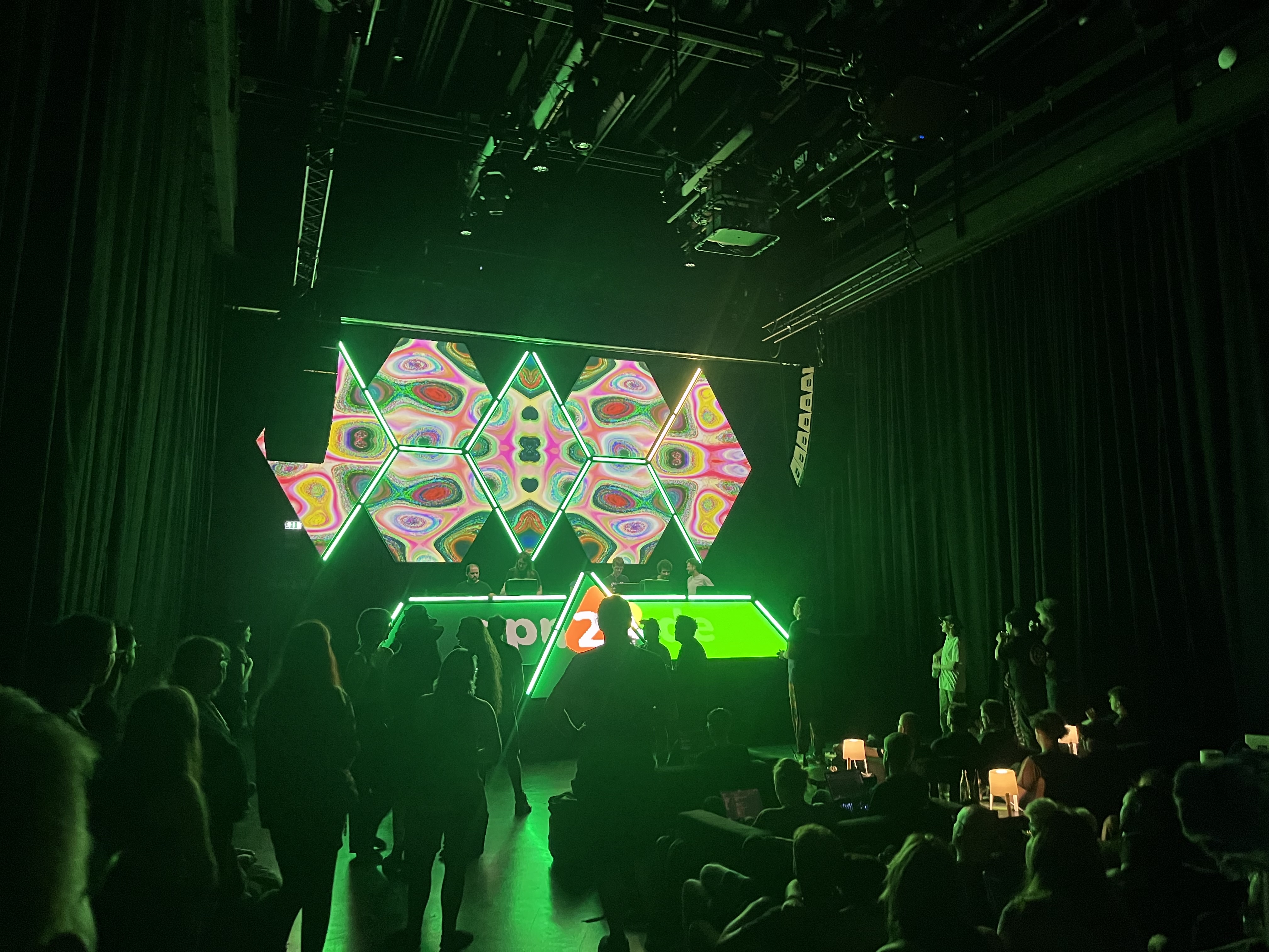 Loading image: a full lounge in the evening, people sitting in the sofas, hacking along on their laptops, talking, drinking tschunk, some dancing in the front before the dj. trippy visuals glaring behind the stage.